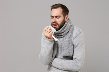 Young man in gray sweater, scarf isolated on grey background. Healthy lifestyle, ill sick disease treatment cold season concept. Mock up copy space. Coughing sneezing covering mouth with paper napkin.