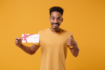 Cheerful young african american guy in casual t-shirt posing isolated on yellow orange wall background in studio. People lifestyle concept. Mock up copy space. Hold gift certificate showing thumb up.