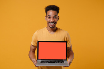 Cheerful young african american guy in casual t-shirt posing isolated on yellow orange wall background. People lifestyle concept. Mock up copy space. Hold laptop pc computer with blank empty screen.