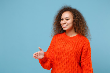 Smiling young african american girl in casual orange knitted clothes isolated on pastel blue background. People lifestyle concept. Mock up copy space. Standing with outstretched hand for greeting.
