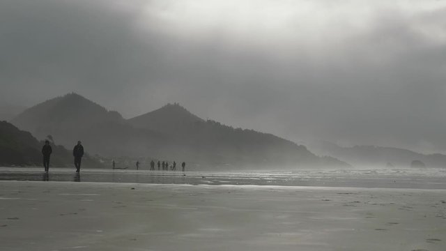 People walking across sand beach to left at Cannon Beach Oregon on a moody overcast cloudy day in mist with mountains in background slow motion 
