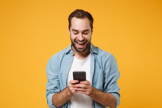 Cheerful Young Man In Casual Blue Shirt Posing Isolated On Yellow Orange Wall Background Studio Portrait. People Emotions Lifestyle Concept. Mock Up Copy Space. Using Mobile Phone, Typing Sms Message.