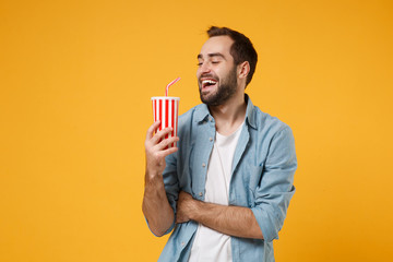 Cheerful young man in casual blue shirt posing isolated on yellow orange wall background, studio portrait. People sincere emotions lifestyle concept. Mock up copy space. Holding cup of soda or cola.