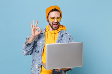 Fototapeta Funny young hipster guy in fashion jeans denim clothes posing isolated on pastel blue wall background. People lifestyle concept. Mock up copy space. Working on laptop pc computer, showing OK gesture. obraz