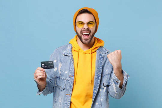 Joyful young hipster guy in fashion jeans denim clothes posing isolated on pastel blue background in studio. People lifestyle concept. Mock up copy space. Hold credit bank card, doing winner gesture.
