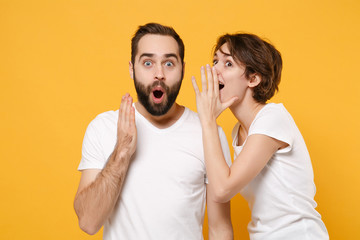 Shocked couple friends bearded guy girl in white t-shirts posing isolated on yellow orange background. People lifestyle concept. Mock up copy space. Whispers gossip and tells secret with hand gesture.