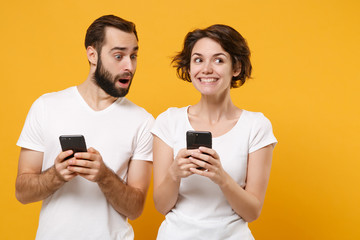 Surprised young couple friends bearded guy girl in white blank empty t-shirts isolated on yellow orange background. People lifestyle concept. Mock up copy space. Using mobile phone typing sms message.