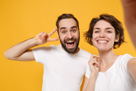 Close Up Of Couple Friends Bearded Guy Girl In White T-shirts Isolated On Yellow Orange Background. People Lifestyle Concept. Mock Up Copy Space Doing Selfie Shot On Mobile Phone Showing Victory Sign.