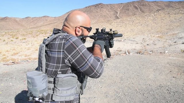 Bald man with a beard firing an assault rifle at an outdoor shooting range in the desert. 24fps.