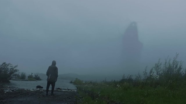 Lonely person watching as a fantastic mythical creature, a giant monster, spectre or a troll walks along the lake and vanishes slowly in the fog. Evening, misty Autumn day.