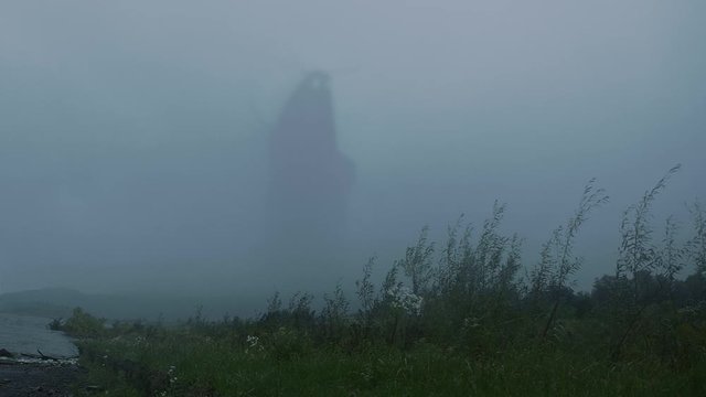 Fantastic mythical creature, a giant monster, spectre or a troll walking along the lake, vanishing slowly in the fog. Evening, misty Autumn day.