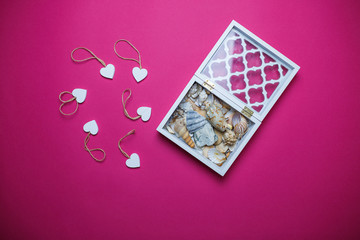 Small white wooden hearts and white wooden box with seashells inside on pink background with copy space. Saint Valentine's day card on pink background.