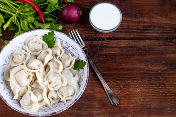 Boiled dumplings with feathers of green onions. In the background are greens, red peppers and bay leaves. In a transparent bowl sour cream.