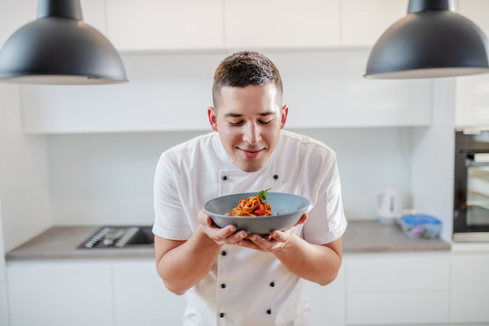 Portrait of handsome satisfied Caucasian chef in uniform standing in kitchen and smelling fresh tagliatelle on plate. Italian cuisine. - Powered by Adobe