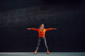 Full length of fit attractive caucasian woman in sportswear and with curly hair standing wide-legged and doing warm up exercises in front of black wall.