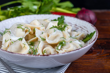 Boiled dumplings with feathers of green onions. In the background are greens, red peppers and bay leaves. In a transparent bowl sour cream.