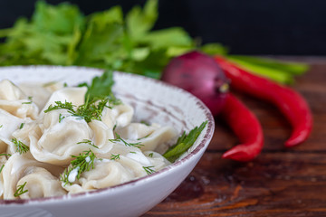 Boiled dumplings with feathers of green onions. In the background are greens, red peppers and bay leaves. In a transparent bowl sour cream.