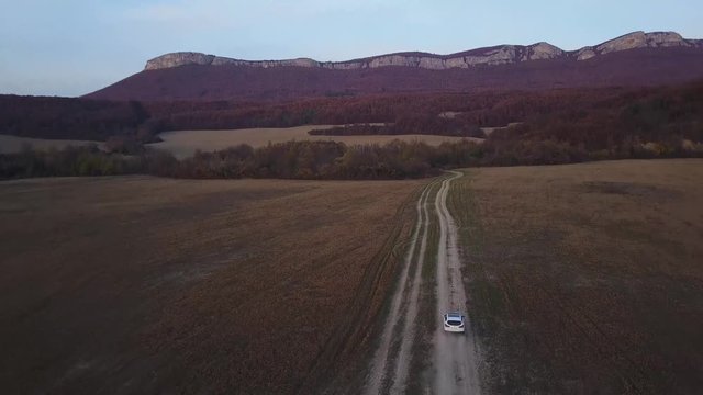Aerial pan of a white car on a dirt road driving towards the mountain at sunset