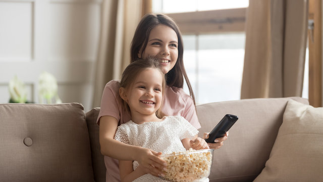 Happy mother with cute little daughter watching tv, eating popcorn