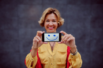 Attractive smiling caucasian blonde senior woman holding smart phone with smiley face on it. Selective focus on hands and phone.