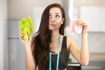 young fit woman with centimeter round her neck holding fresh salad in one hand and slice of ham in another looks doubtful,nutrition and dietology