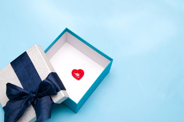 red heart figurine with a smile in a gift box with a large dark blue satin ribbon bow, white box inside, blue background, copy space