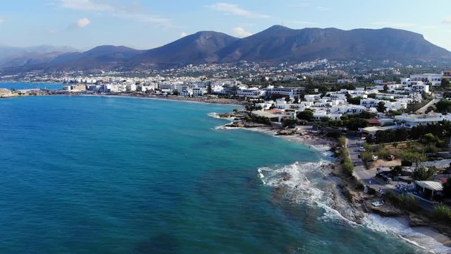 Tourist town at sea shore, many buildings at waterfront, rough stony beach, tall mountains on backdrop. Aerial panorama of Chersonisou area, north coast of Crete Island