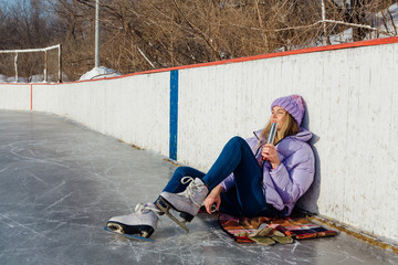 Lovely young woman relaxing after riding ice skates and drinking hot drink from termo pot on the ice rink.