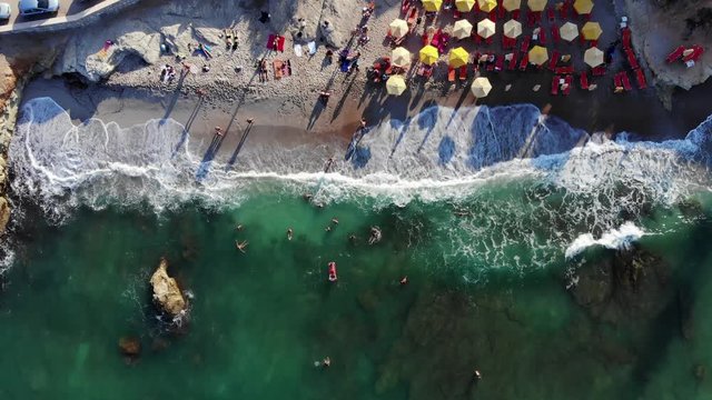 People rest at small beach in evening time, top-down aerial shot. Half area with loungers and umbrellas, other part is empty sand. Waves wash and seethe with white foam at shallow rocky sea bottom