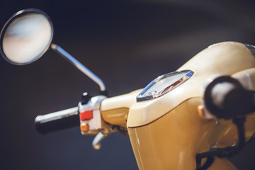 The steering wheel of a vintage beige moped with a dashboard and a round rearview mirror is illuminated with bright light.