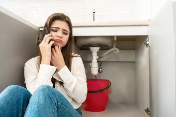 young beautiful woman looks desperate facing leaking pipe consulting specialist on the phone