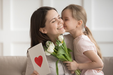 Little daughter kissing mother on cheek, presenting flowers and gift