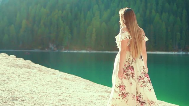 mysterious woman casual dress walks on white shore alpine lake, goes to sun rays enjoy nature. Backdrop forest trees turquoise water. Slow motion shoot back lady without face, turned away. long hair