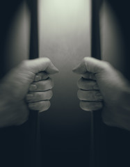 In jail concept with male hands holding jail bars, light in background room, first person view, toned image