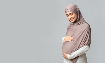 Pregnant muslim woman embracing her belly, posing over grey background