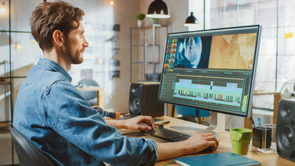 Fototapeta premium Beautiful Creative Male Video Editor with Beard and Jeans Shirt Works with Footage on His Personal Computer with Big Display. He Works in a Cool Bright Office Loft.