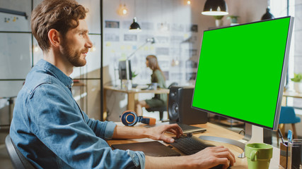 Obraz premium Male Creative Designer with Beard and Jeans Shirt Works on His Personal Computer with Big Green Screen Mock Up Display. He Works in Cool Office Loft. Female Creative Colleague Works in Background.
