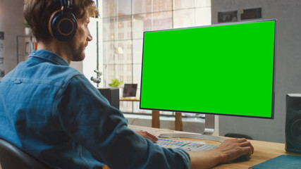 Young Professional Creative Employee Works on His Personal Computer with Big Green Screen Mock Up Display. He Works in a Cool Office Loft. Big Window with City View in the Background.