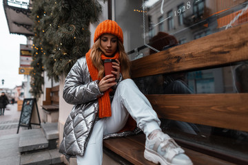 Cute young woman in an orange knitted trendy hat with a vintage scarf in a stylish silver jacket in...