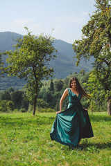Fototapeta premium Happy, brunette girl is played with a dress. Carpathians mountains at the background. Young romantic woman in long, silk, green dress on the mountainside.