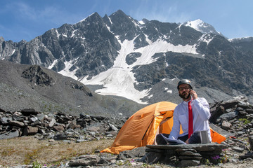Outdoor office. Office worker in white shirt, red tie and climbing helmet works in alpine landscape.