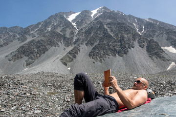 A mountain climber lies topless on a stone and reads a book in alpine landscape.