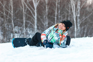 Fototapeta na wymiar Beautiful young blond woman snowboarder in pigtails in colorful costume is smiling, sitting on top of the mountain. Winter sunny day