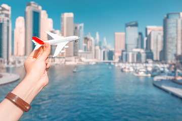 A hand holds a toy white passenger plane against a backdrop of skyscrapers and a seaport. Concept of travel and ticket prices