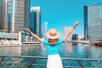 A happy tourist girl holds up her hands and enjoys the stunning view of the skyscrapers in the Dubai Marina area. Travel to the United Arab Emirates