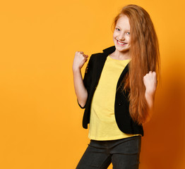Ginger schoolgirl in black jacket, pants, yellow t-shirt. She is smiling, looking satisfied, posing on orange background. Close up