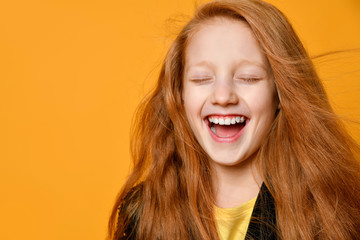 Red-headed teenage child in black jacket and yellow t-shirt. She laughing with closed eyes, posing on orange background. Close up