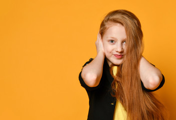 Red-headed teenage kid in black jacket and yellow t-shirt. She closed her ears with hands, posing on orange background. Close up