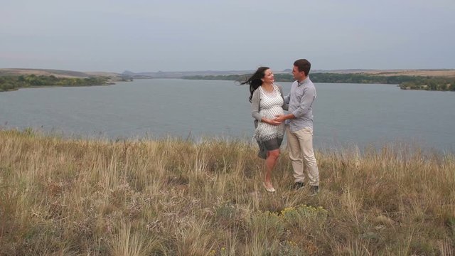 A happy man gently strokes the tummy of a pregnant wife standing in a field against the background of a large lake in windy weather.