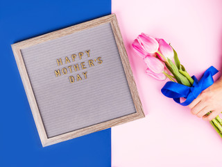 Mother's day greetings on letter board and female hands with red manicure holding beautiful tulips on pink and classic blue trendy split backdrop.